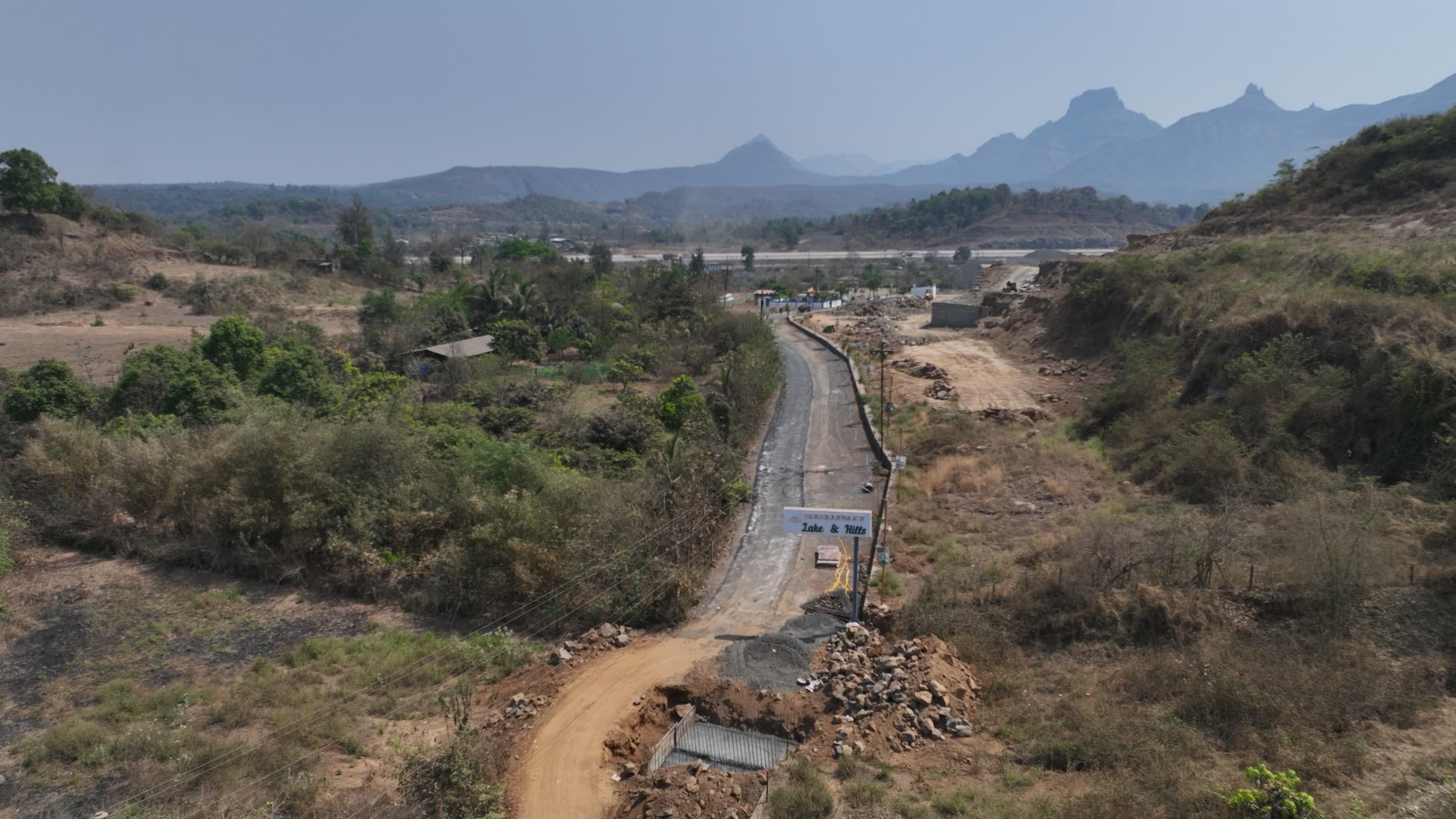 Low-altitude photograph looking along the approach road into the site, with the Varchaswaa's Lake & Hills signboard at the roadside and the hill range on the horizon.